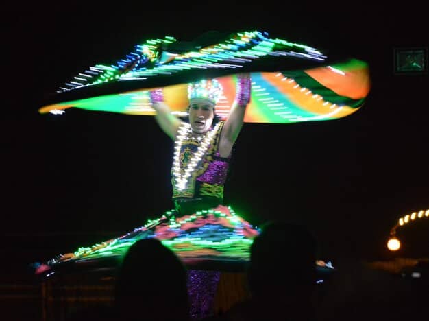 A futuristic image showing a Rio Carnival parade float with holographic projections, where performers seem to interact with the digital elements. The background showcases spectators wearing augmented reality glasses, indicating an enhanced viewing experience through technology.